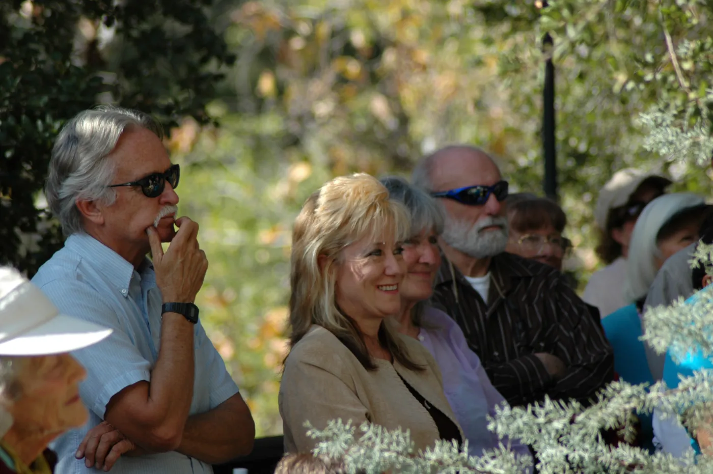 A group of people in a tree grove, all looking off camera at something. Two men and two women are in focus. The two men have their arms are crossed and the two women are smiling.