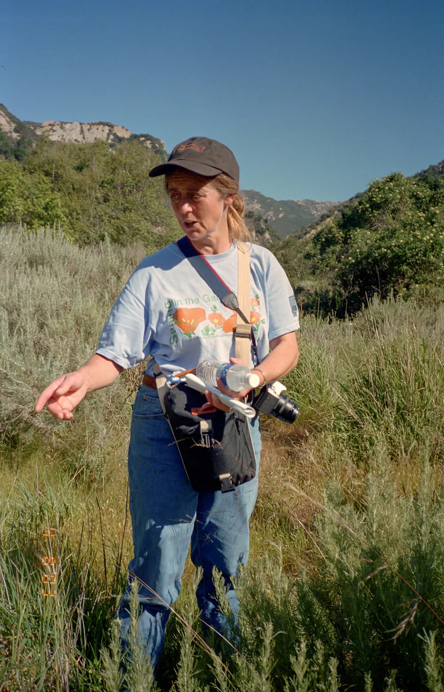 A color photo of a woman in a cap and t-shirt standing in a grassy field with a sky above, pointing with one hand and carrying binoculars and field gear around her waist.