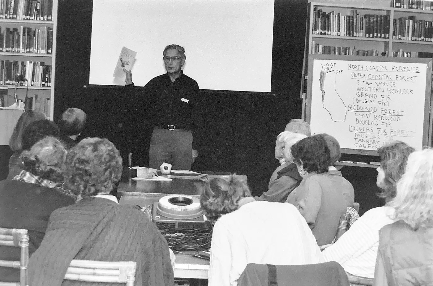 A black and white photo of a man holding up a piece of paper in front of a group of people sitting at a table with a whiteboard to the right.