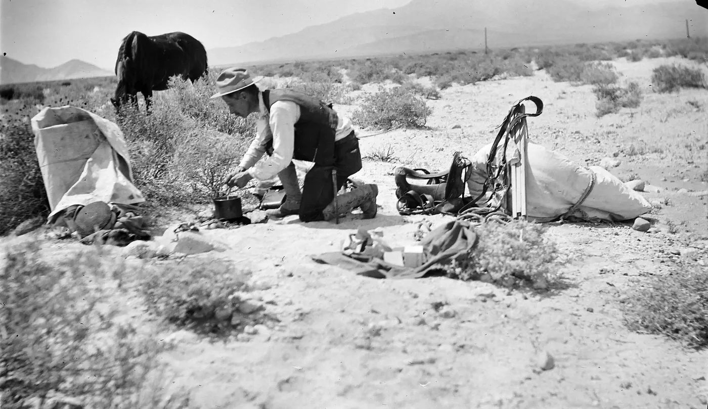 a black and white photo of a man kneeling on desert ground stirring a pot over a small fire with packs and gear spread nearby and a horse grazing in the background.