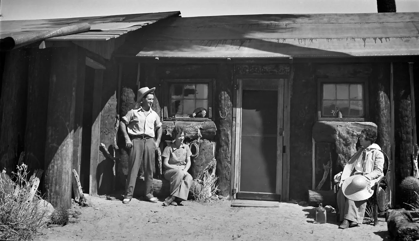 A black and white photo of a man in a cowboy hat standing and two women sitting outside an old wooden building. 
