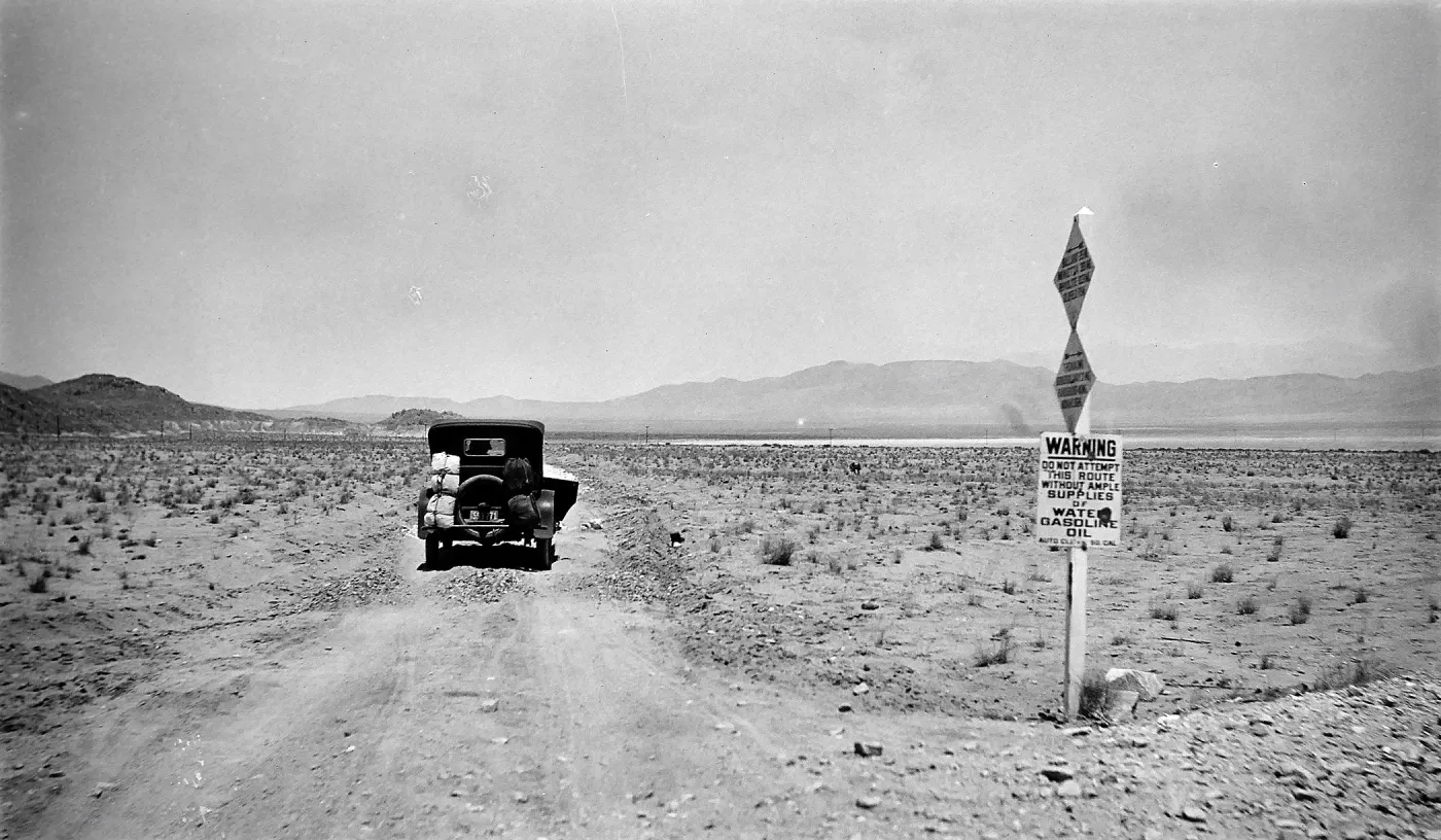 Black and white photograph of a black car driving down a dirt road into the desert past a sign that says Warning.