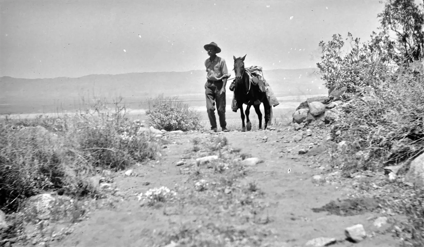 Black and white photograph of a man in a hat walking a black horse down a rock-strewn desert pathpath.