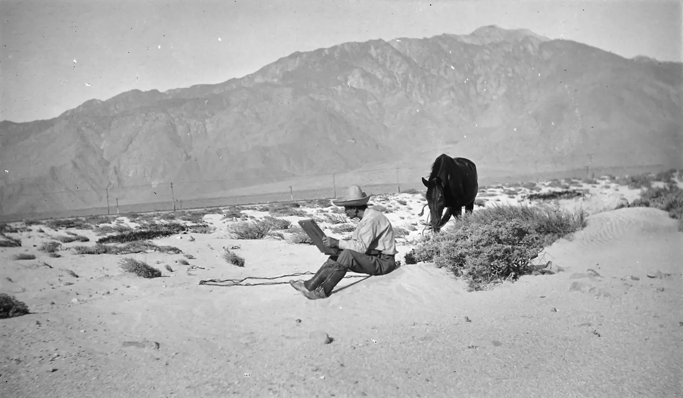 Black and white photo of a man in a hat sitting and sketching in a large book with a black horse next to him and a mountain range behind him.