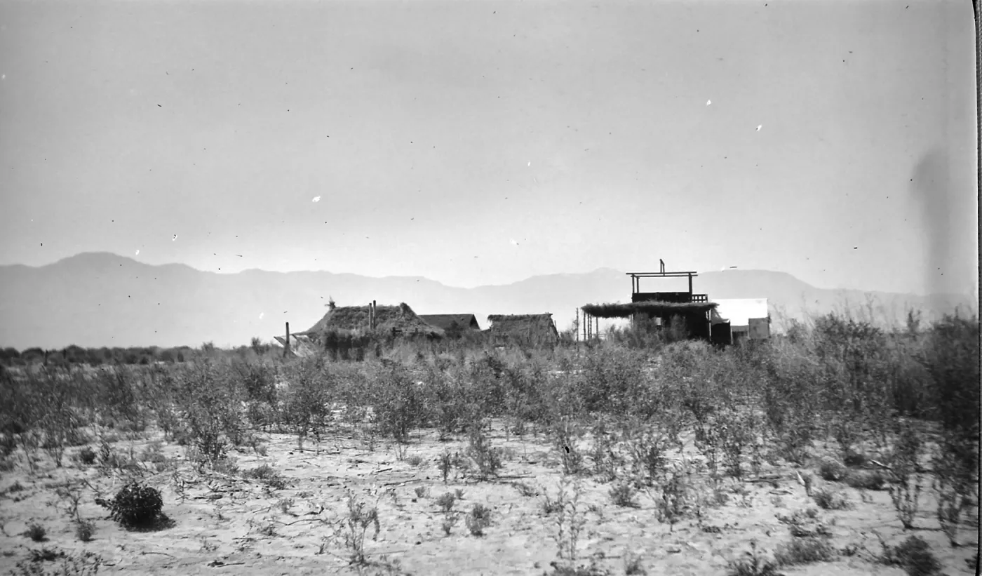Black and white photograph of a group of buildings in a desert setting with brush in the foreground and hills in the distance.