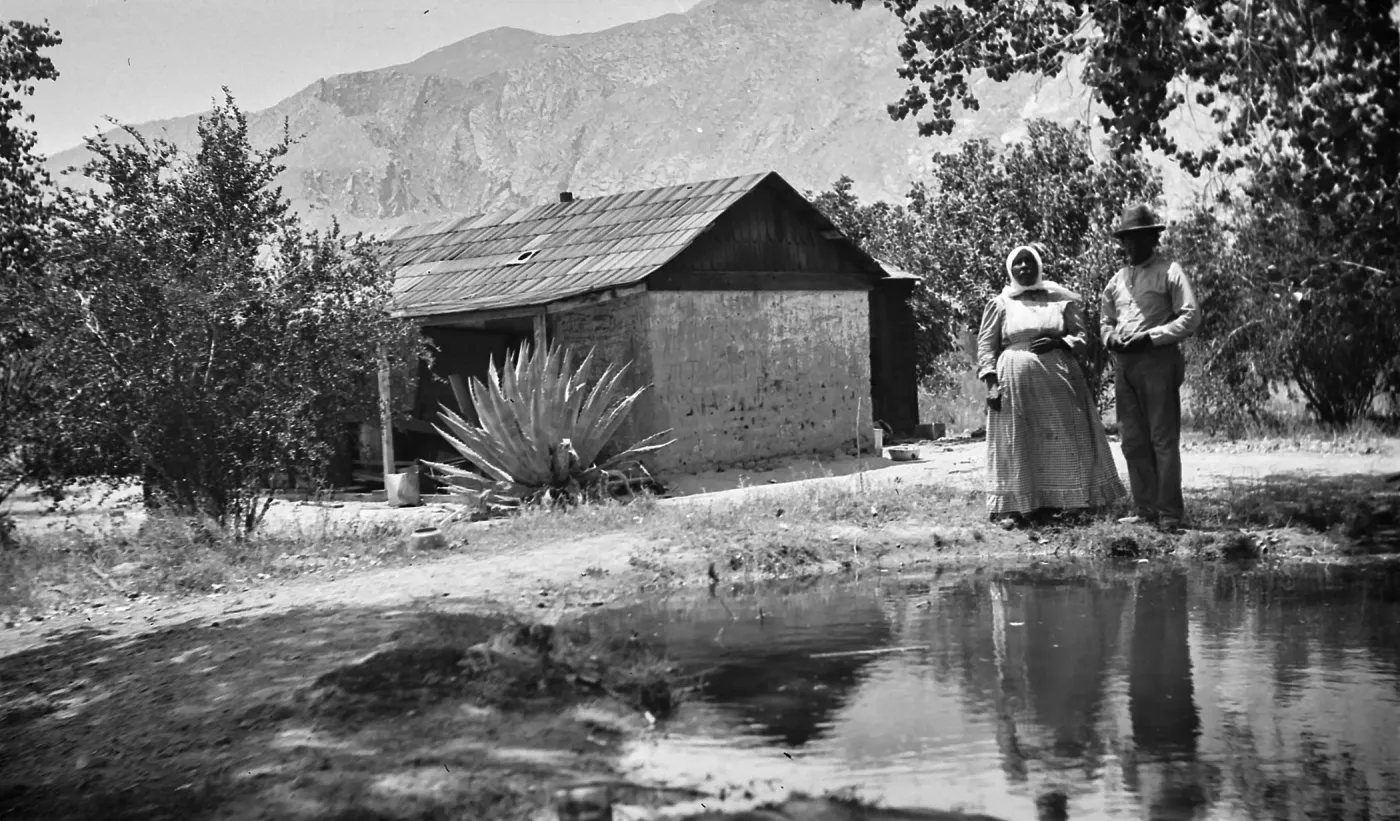 Black and white photograph of a man in a hat and a woman in a dress and head covering next to a pool of water, with a wooden house, trees, desert plants, and mountains in the background.