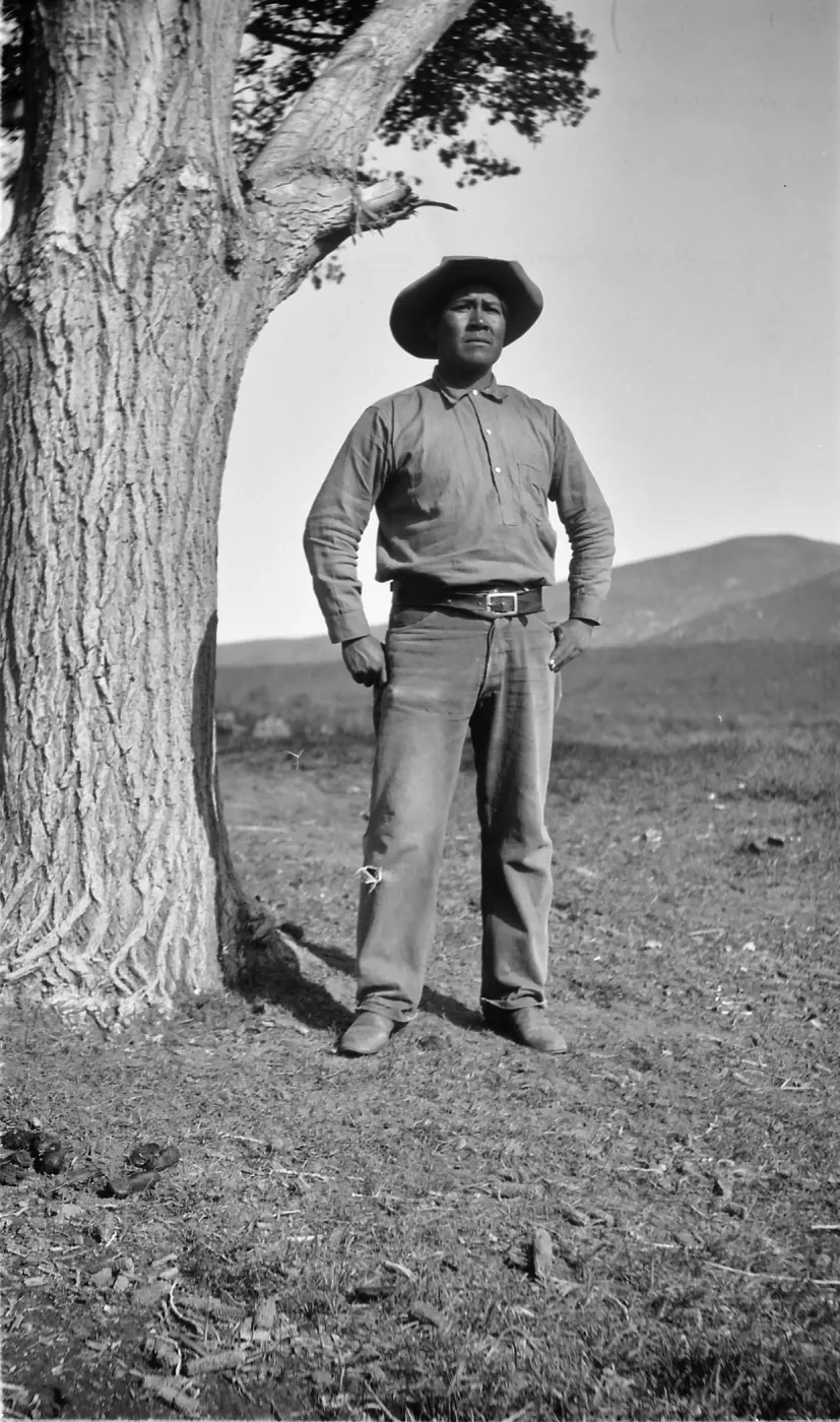 Black and white photograph of a man in a hat, buttoned shirt, and jeans next to a tree with a serious expression.