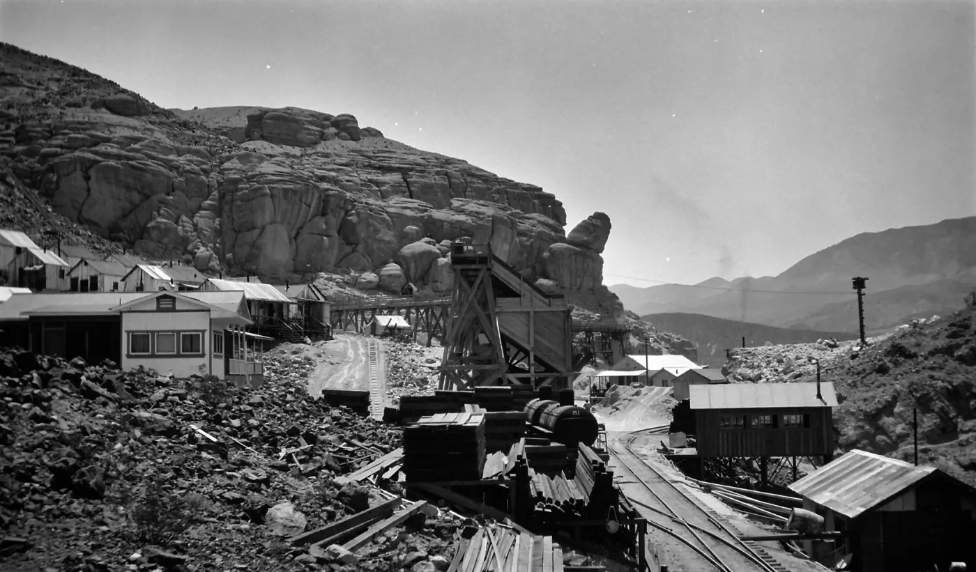 Black and white photograph of train tracks and wooden buildings beside a rocky hill with hills in the distance.