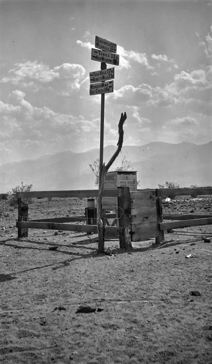 Black and white photograph of a signpost above a wooden fence that has signs to Blackwater, Wildrose, Tanks, Ash Meadow, Tule Hole, Bennetts Wells, Wondermine, and Salt Creek. There are mountains in the distance.