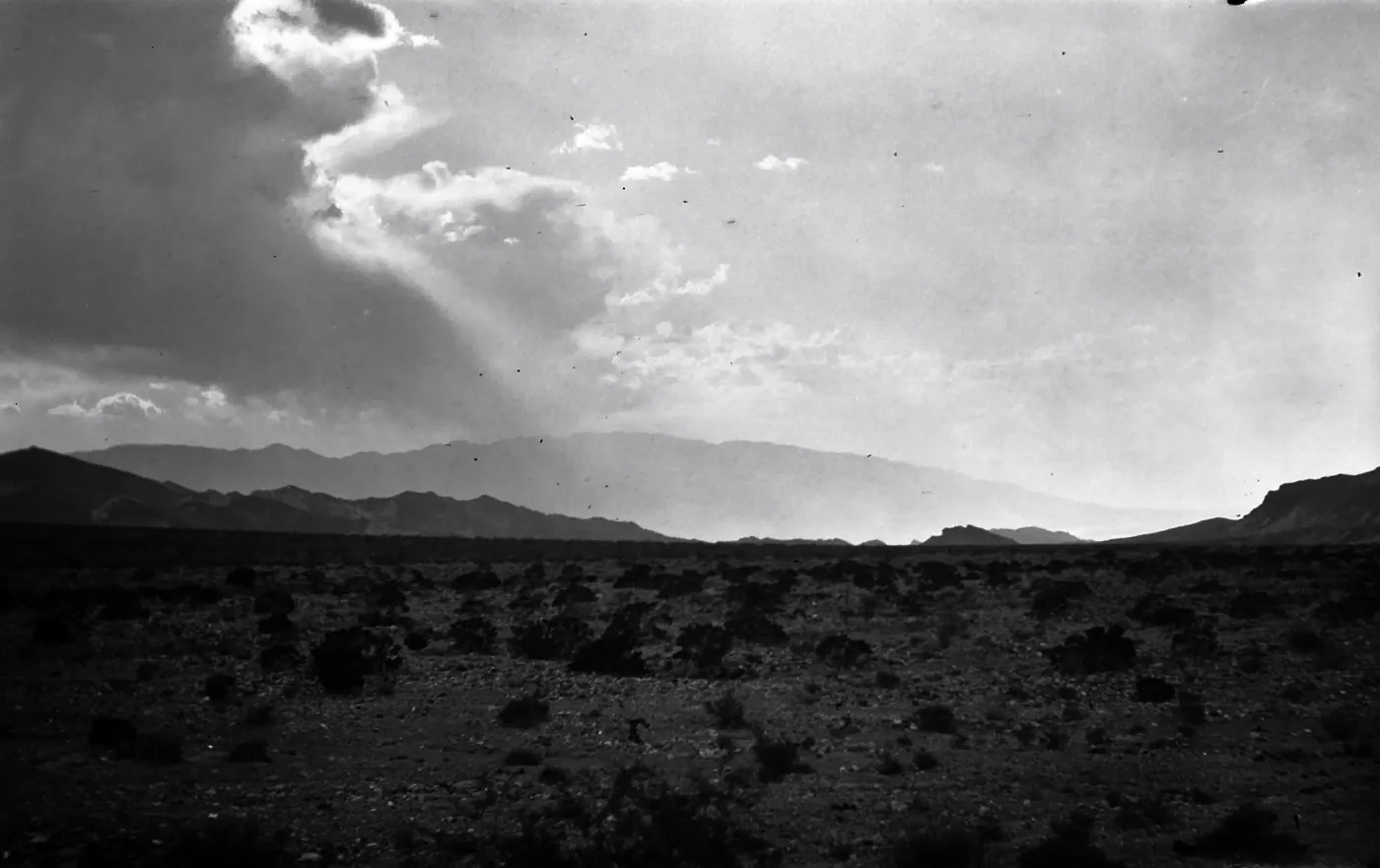 Black and white photograph of a desert landscape with rays of sunlight coming through clouds and mountains in the distance.