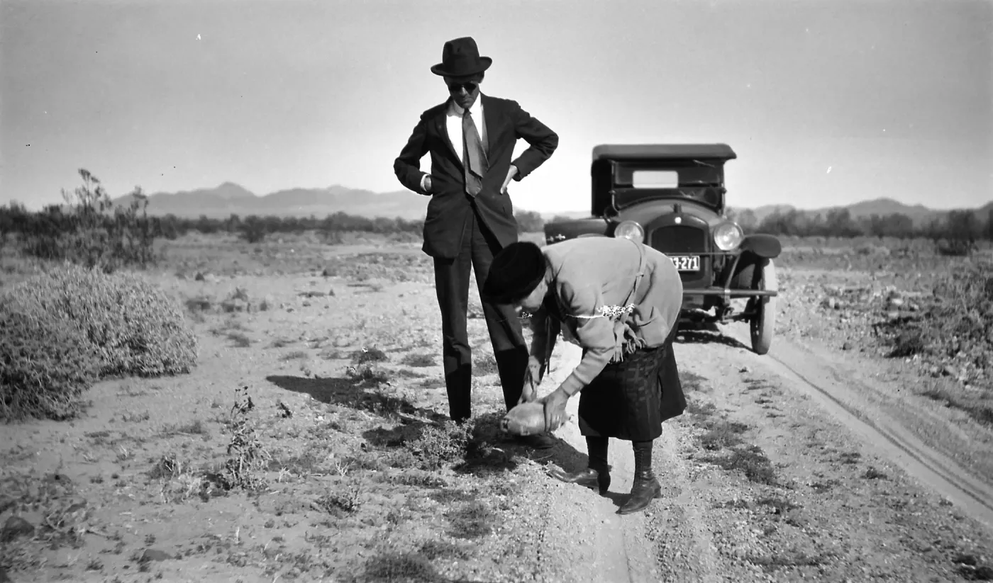 Black and white photograph of a man and woman on a dirt road with a black car behind them. The man is wearing a suit and the woman is lifting a tortoise out of the road.