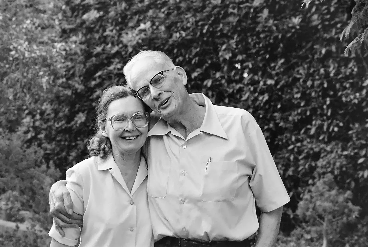 Black and white photograph of a man and woman against a background of bushes. Both are wearing glasses and white shirts and smiling at the camera. 