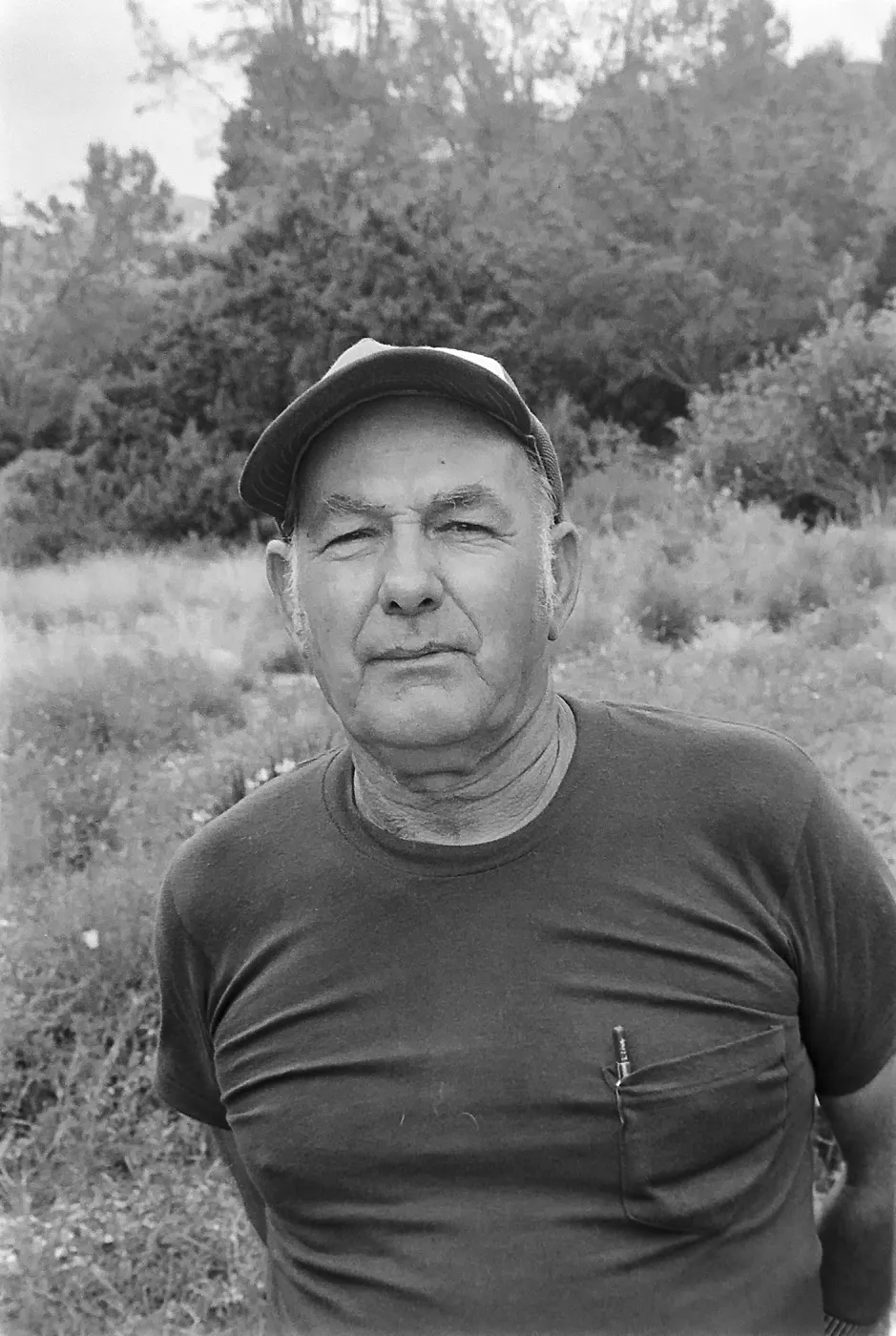 Black and white photograph of a man wearing a short sleeved shirt and a baseball cap, looking at the camera in a garden setting.