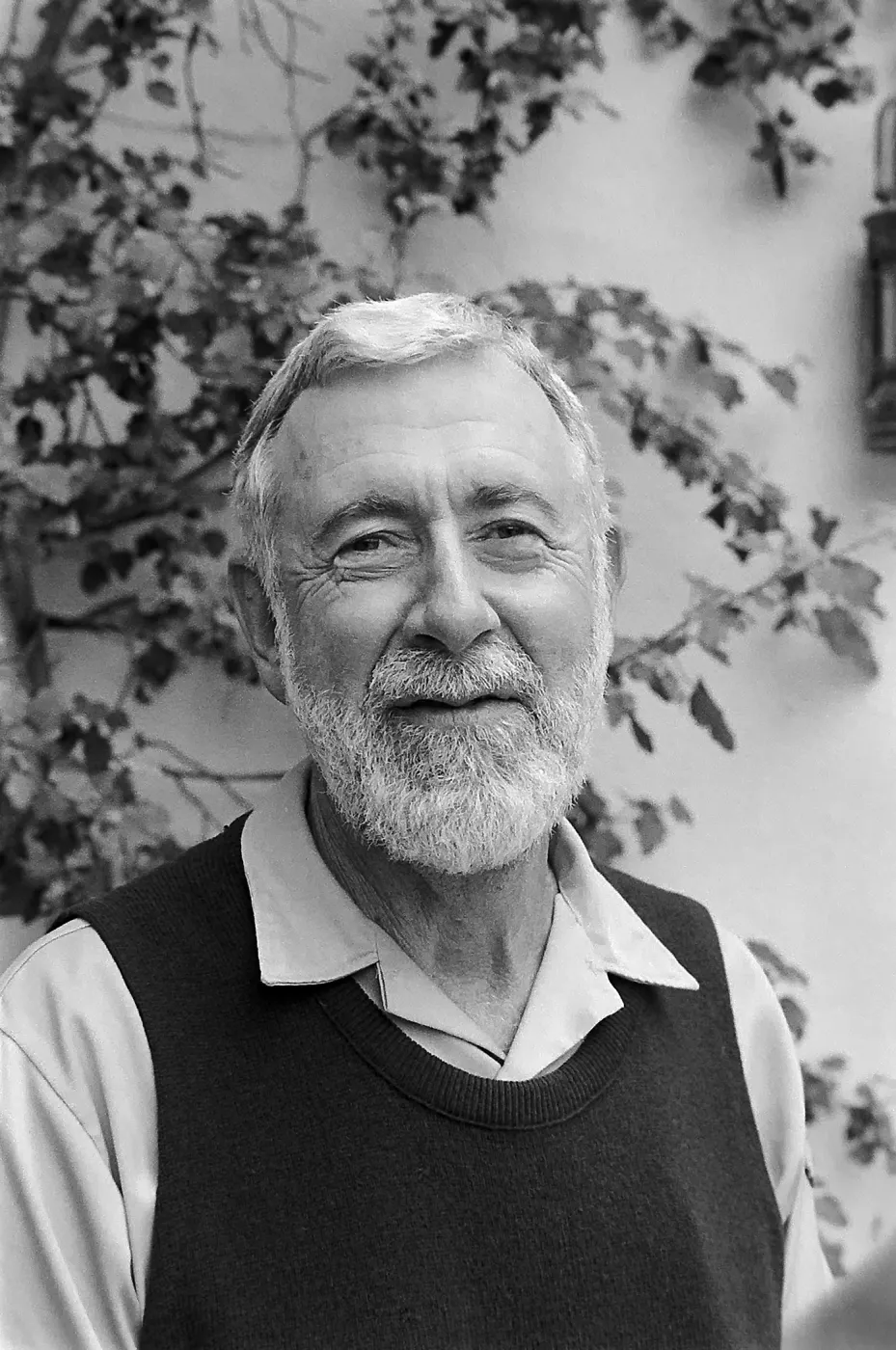 Close-up portrait of an older smiling man with a beard and sweater vest against a leafy wall background.