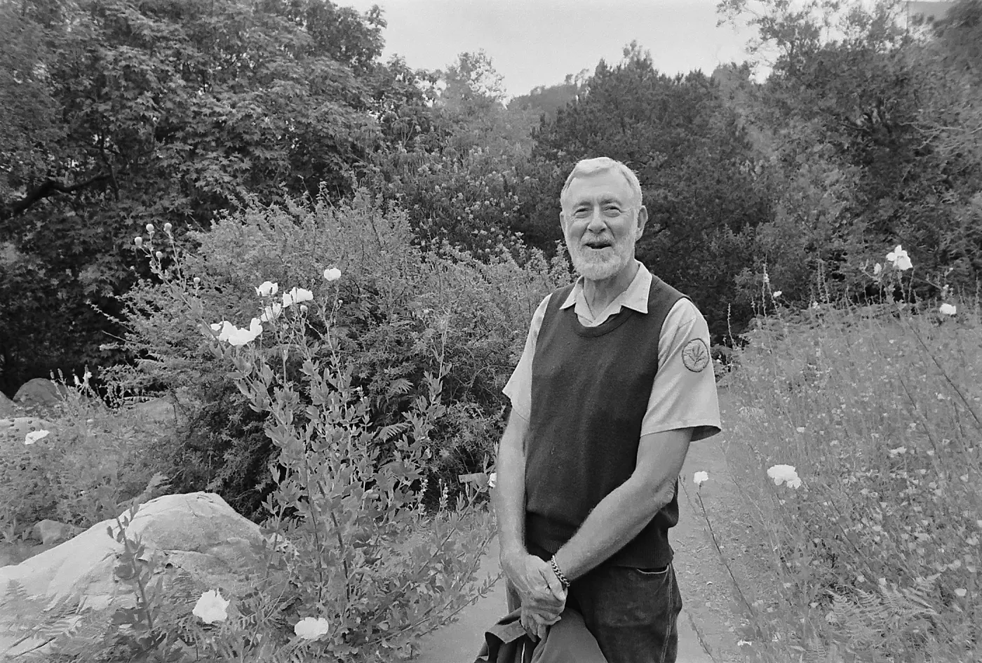 Black-and-white photo of an older man in a vest smiling and standing on a garden path surrounded by wildflowers, shrubs, and trees.