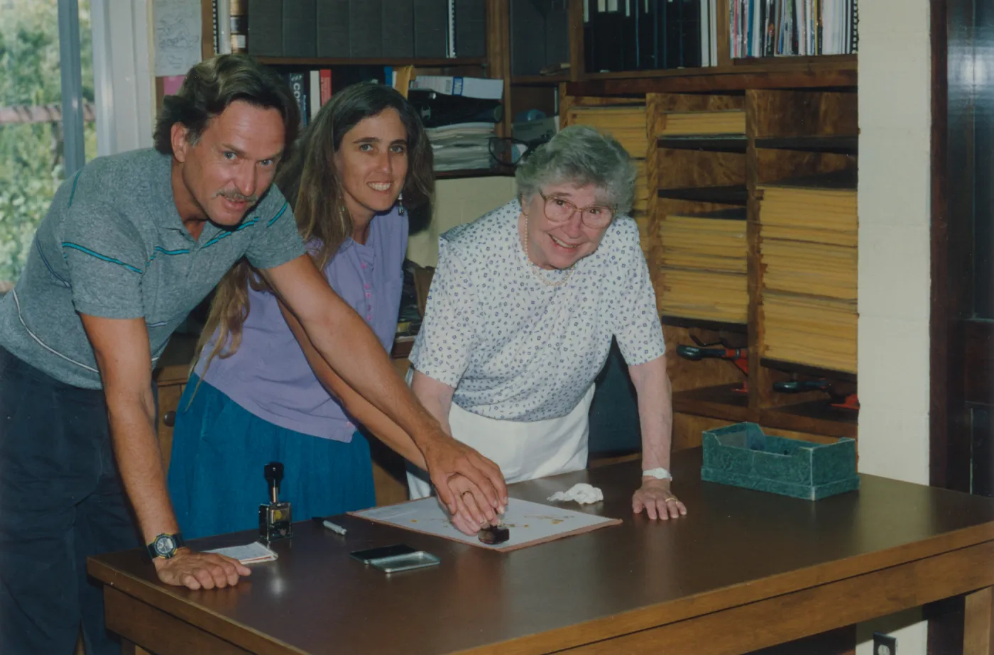 A man in grey, a woman with long hair wearing in a lavender shirt, and an older woman in white look toward the camera while standing in front of shelves lined with many binders, books, and yellow memo pads. The three lean forward over a desk where they put their hands together, applying a red stamp to a document sitting on the desk. .