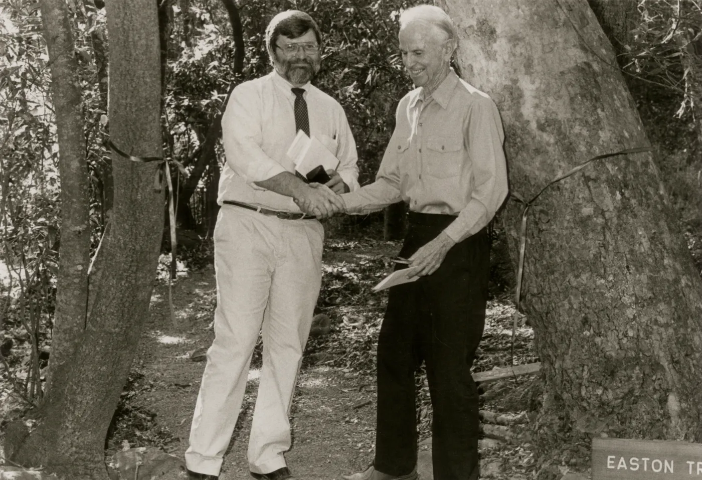 A black and white photo of two men, one is bearded, wearing glasses and a light-colored dress shirt and dark tie, the other has light hair and wears dark slacks. Both are holding documents and stand in front of a cut ceremonial ribbon tied between two trees. A sign reading "Easton Tr" is cutoff at the edge of the image. 