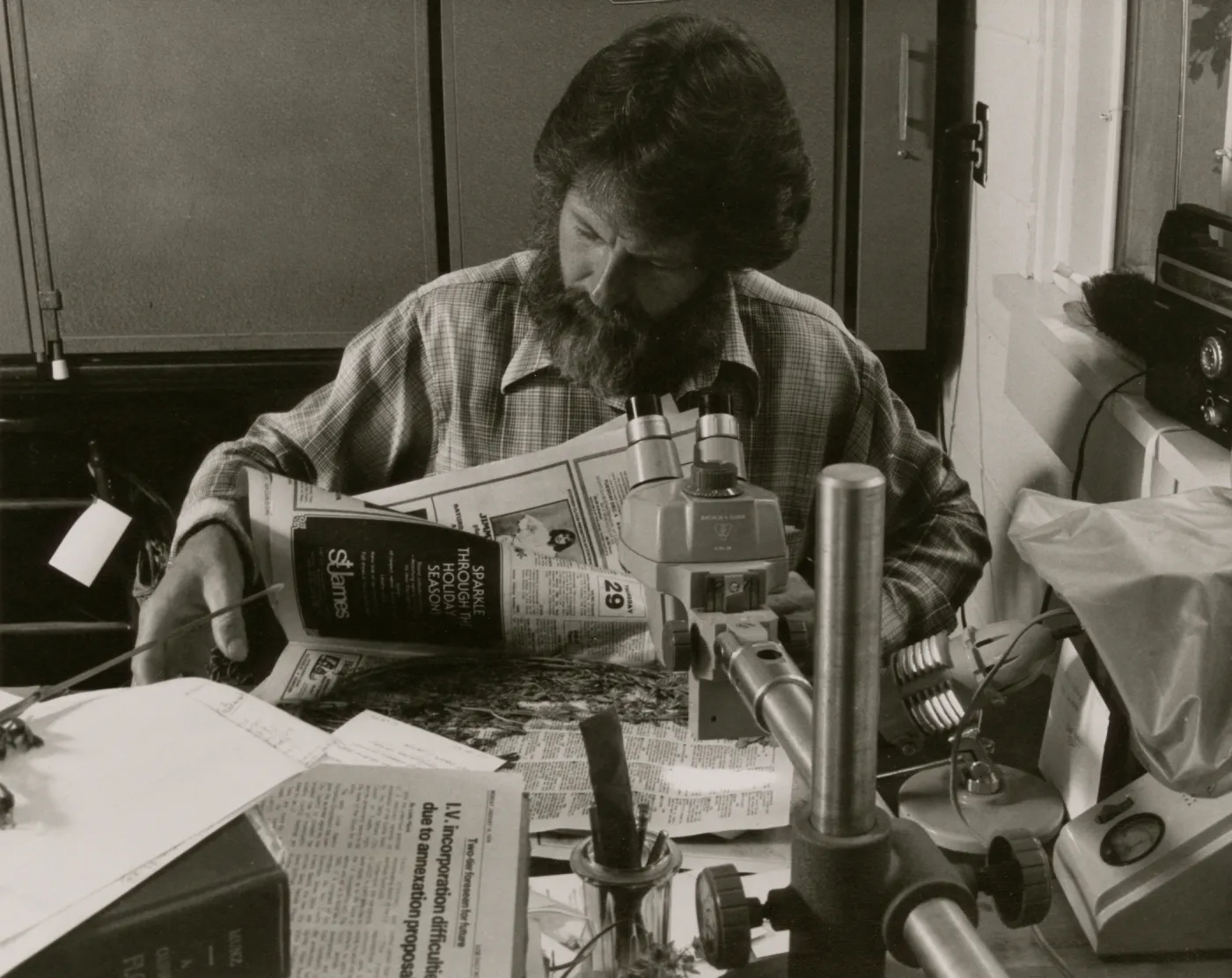 A black and white photo of a bearded man in a plaid shirt looking through newspapers while sitting at a desk cluttered with newspapers, books, and scientific instruments.