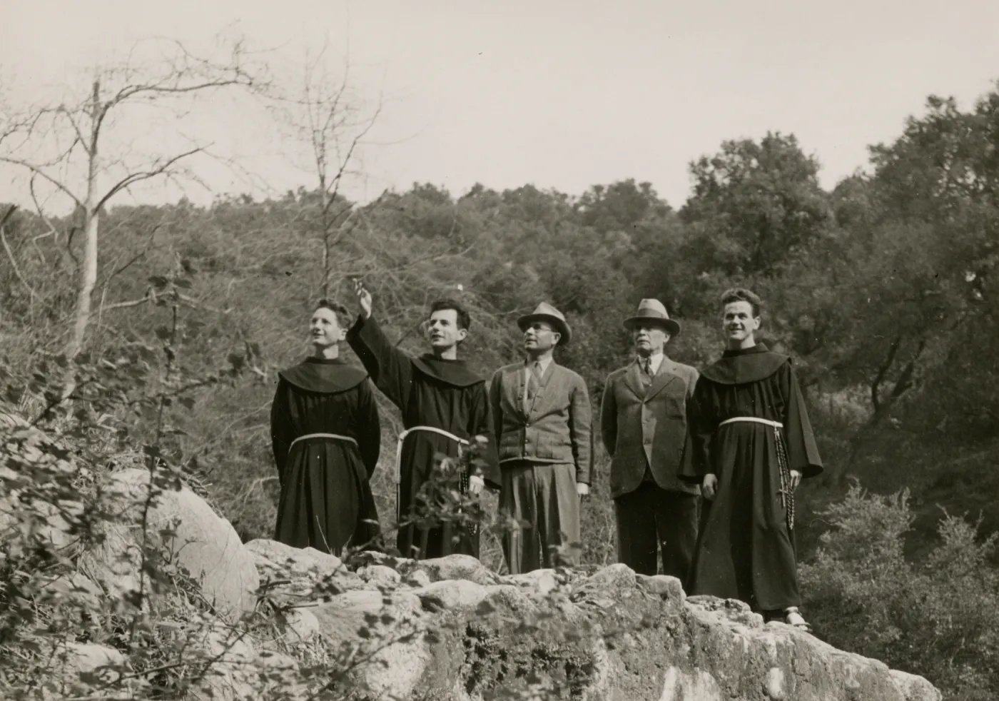 A black and white photo of five men look into the distance while standing on a rocky bluff. Three of the men wear robes ties with ropes at the waist and two wear suits and trilby hats. One of the robbed men points out in the distance where the group is looking.