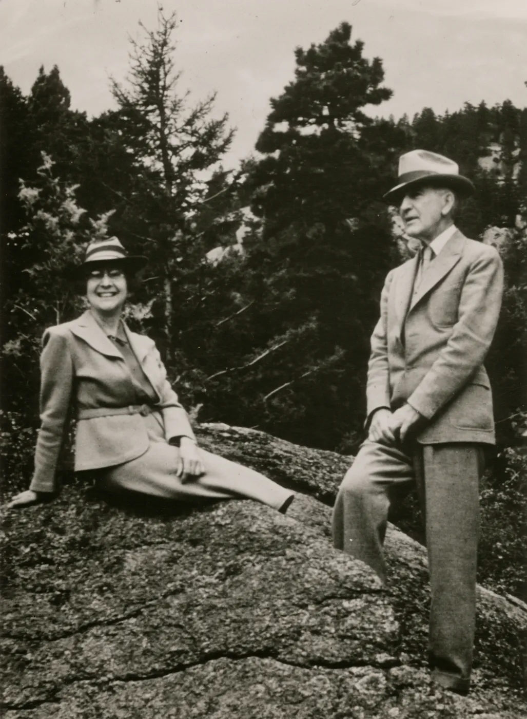 A black and white photo of a man and woman atop a rocky outcropping in front of a forest of evergreen trees. Both wear suits and trilby hats, the woman sits on the outcropping, smiling at the camera, and the man stands to the side looking out toward the distance.