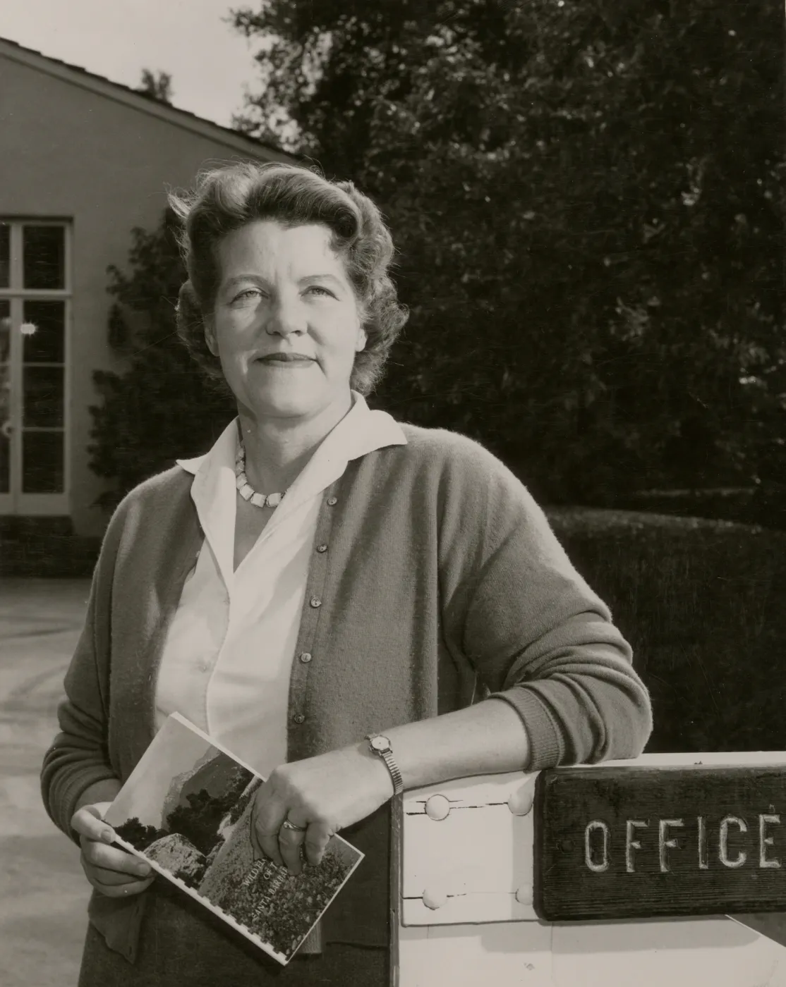 A black and white photo of a woman in a blouse posing in front of a building with neatly trimmed hedges. She holds a booklet depicting a field in front of a forest and mountain and one of her arms rests upon a wooden fence gate with a sign reading "Office" cutoff at the edge of the image.