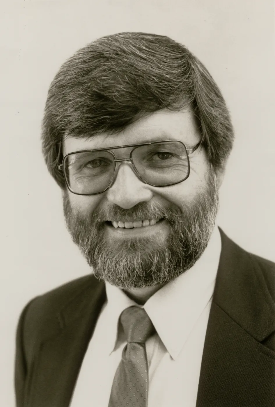 A black and white close-up of a bearded man in a suit and squarish glasses in front of a blank background. 