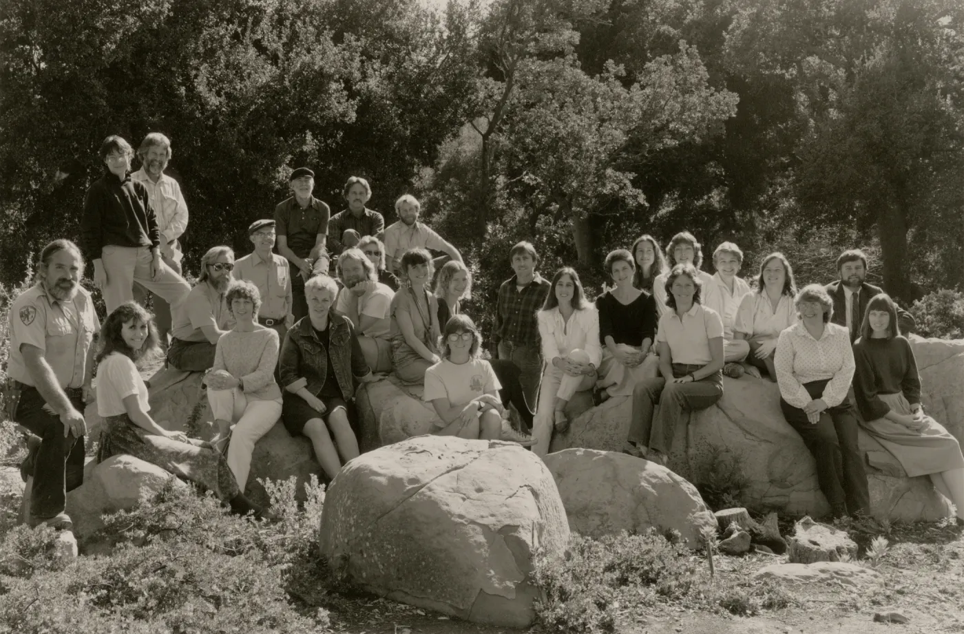 A black and white photo of 27 people are posed sitting atop or standing around several boulders a the edge of a forest.