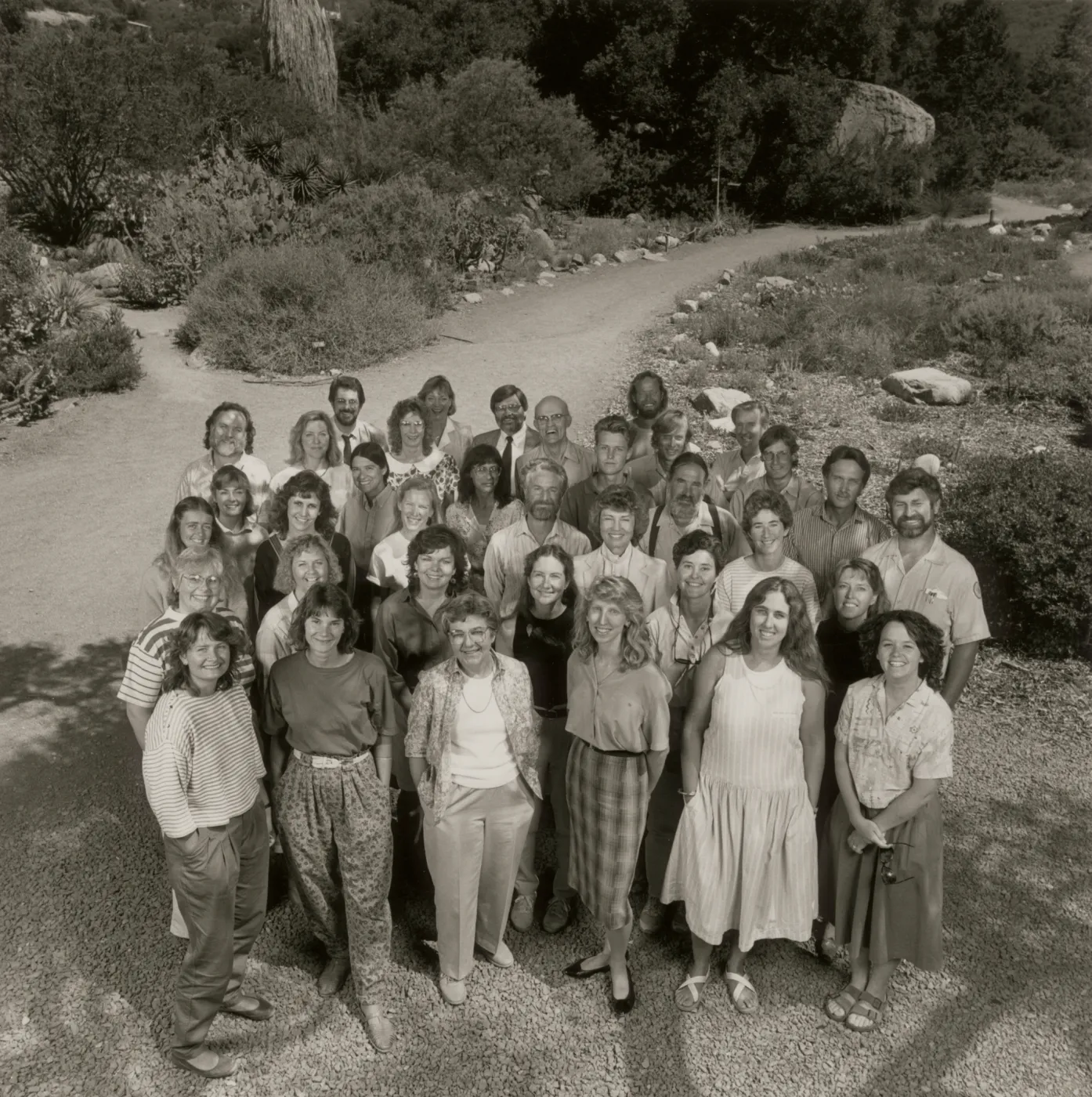 A black and white photo of a group of 36 smiling people taken from a high angle. They stand in front of a gravel road lined with stones and rough shrubbery.