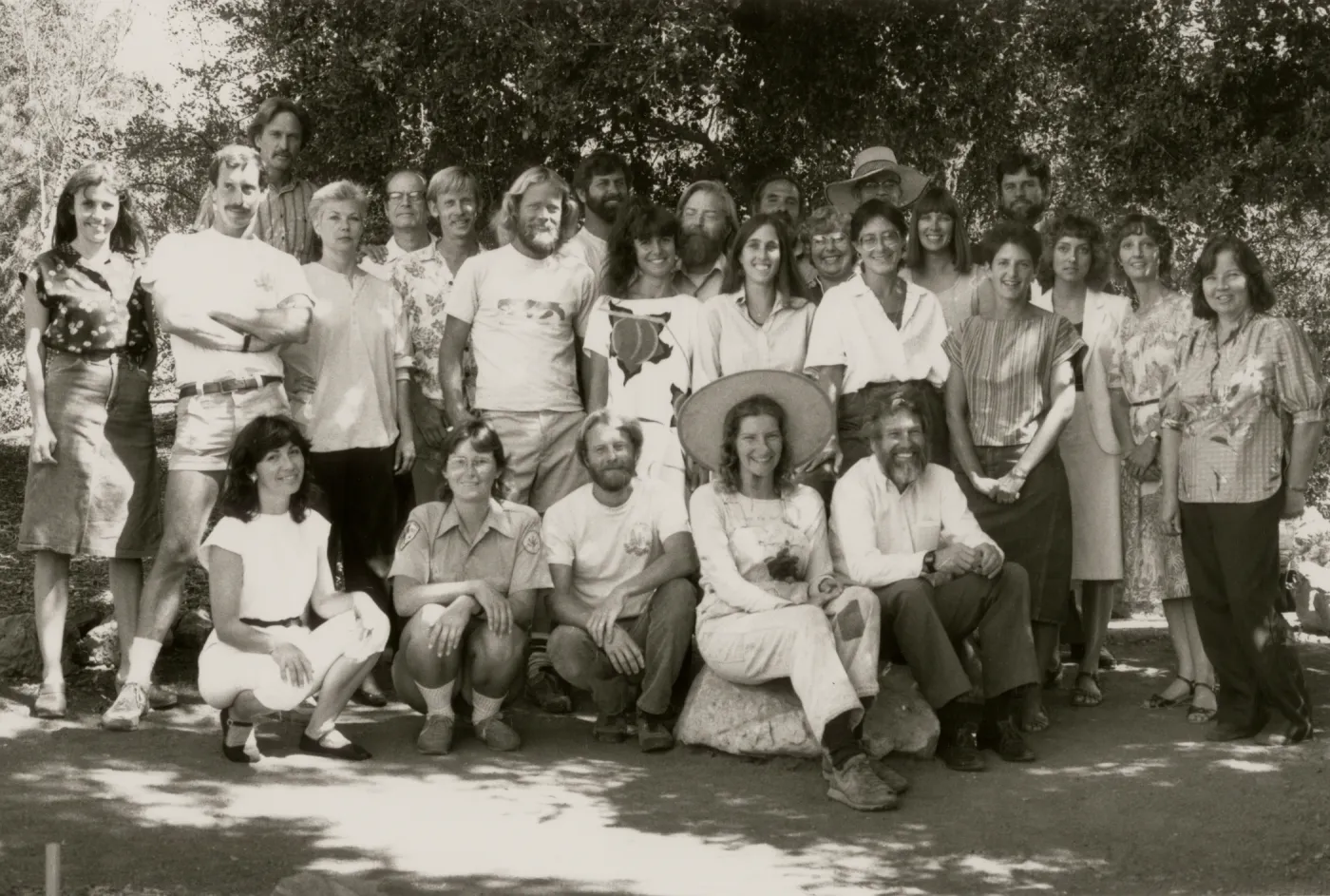 A black and white photo of a group of 26 smiling people posed on the edge of a path. Five people crouch or sit on a large rock, the rest stand in two rows behind them.