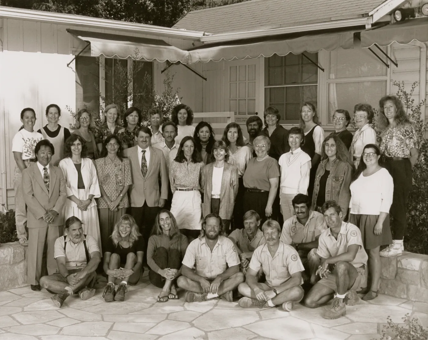 A black and white photo of 34 people posed in the corner of a stone patio in front of a one-story building. Eight people sit or kneel on the ground and the rest are arranged in two rows behind them.
