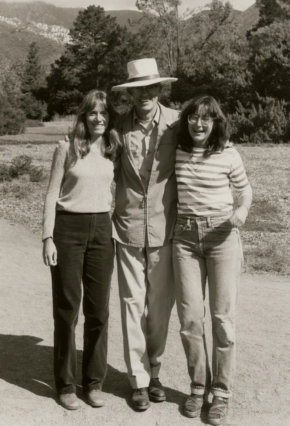 A black and white photo of a smiling man wearing a suit and large, wide-brimmed hat and two smiling women in causal dress in a rocky field in front of a copse of trees.