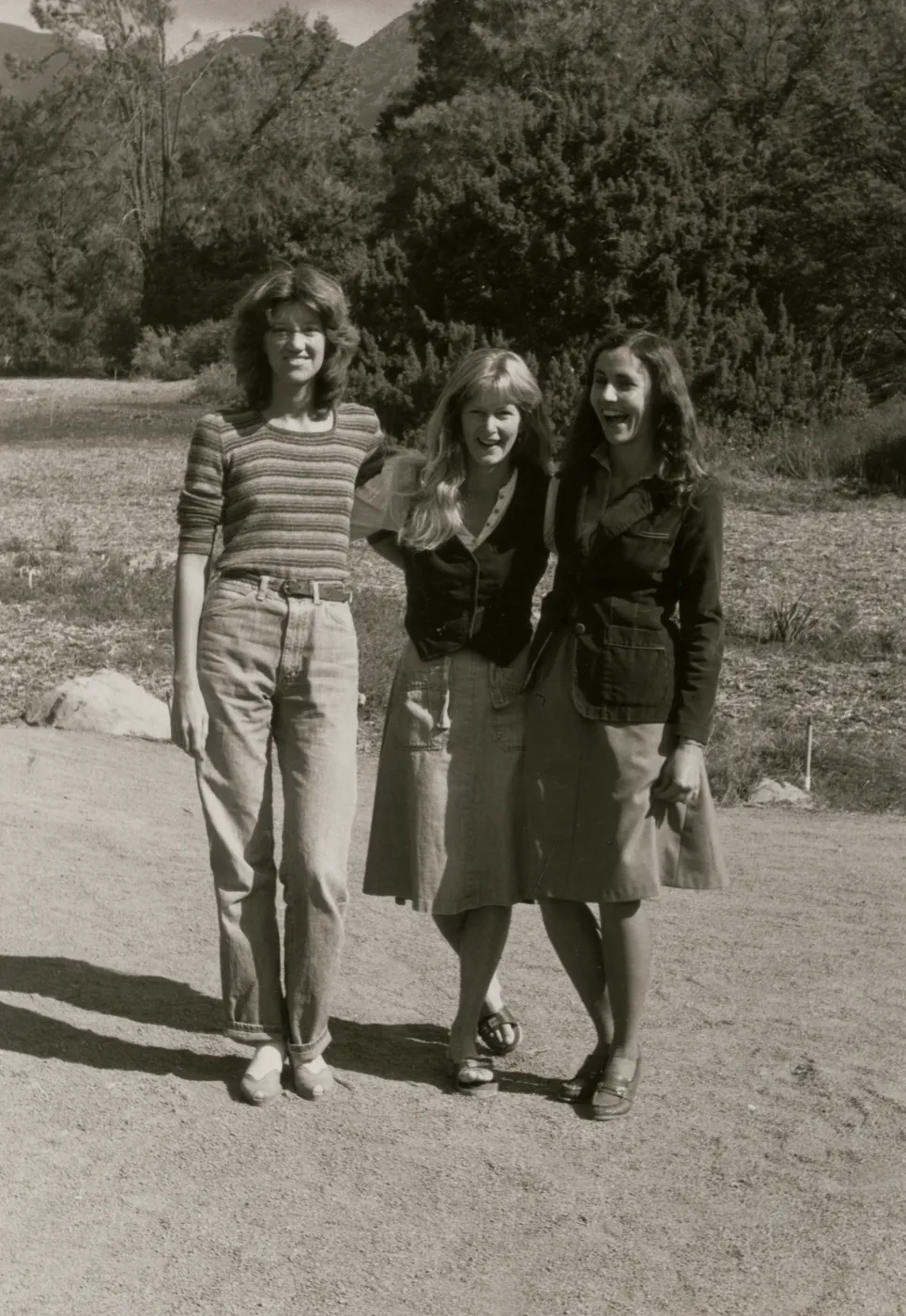A black and white photo of three jovial women with their arms around each other in a rocky field in front of a copse of trees. 