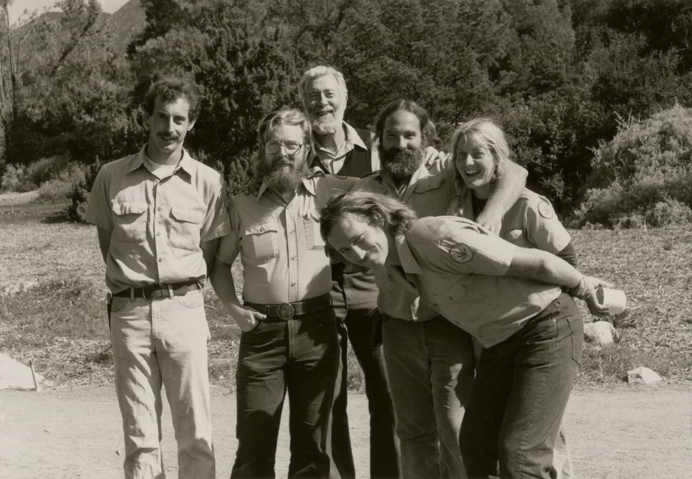 A black and white photo of six smiling people in a rocky field in front of a copse of trees. One man leans in, bent double from the side, in front of the rest of the group.