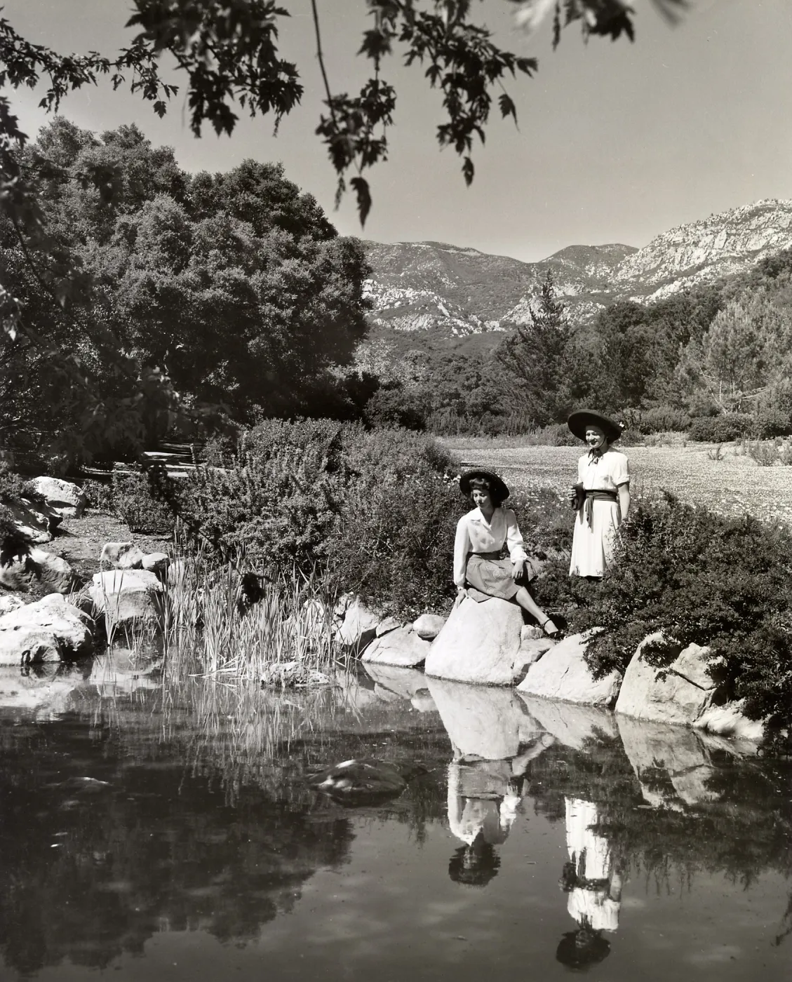 Two young women enjoy their own reflections in a quiet pool with a background of sand strawberries in bloom and mountains rising above