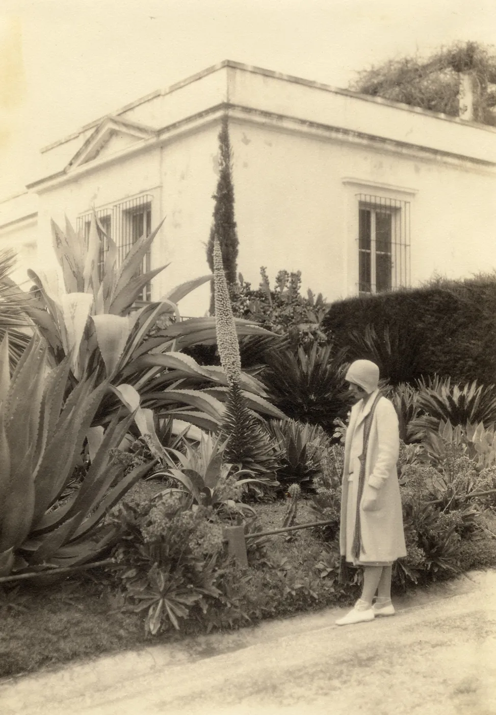 Black and white photograph of woman looking a plant bed in front of white building.