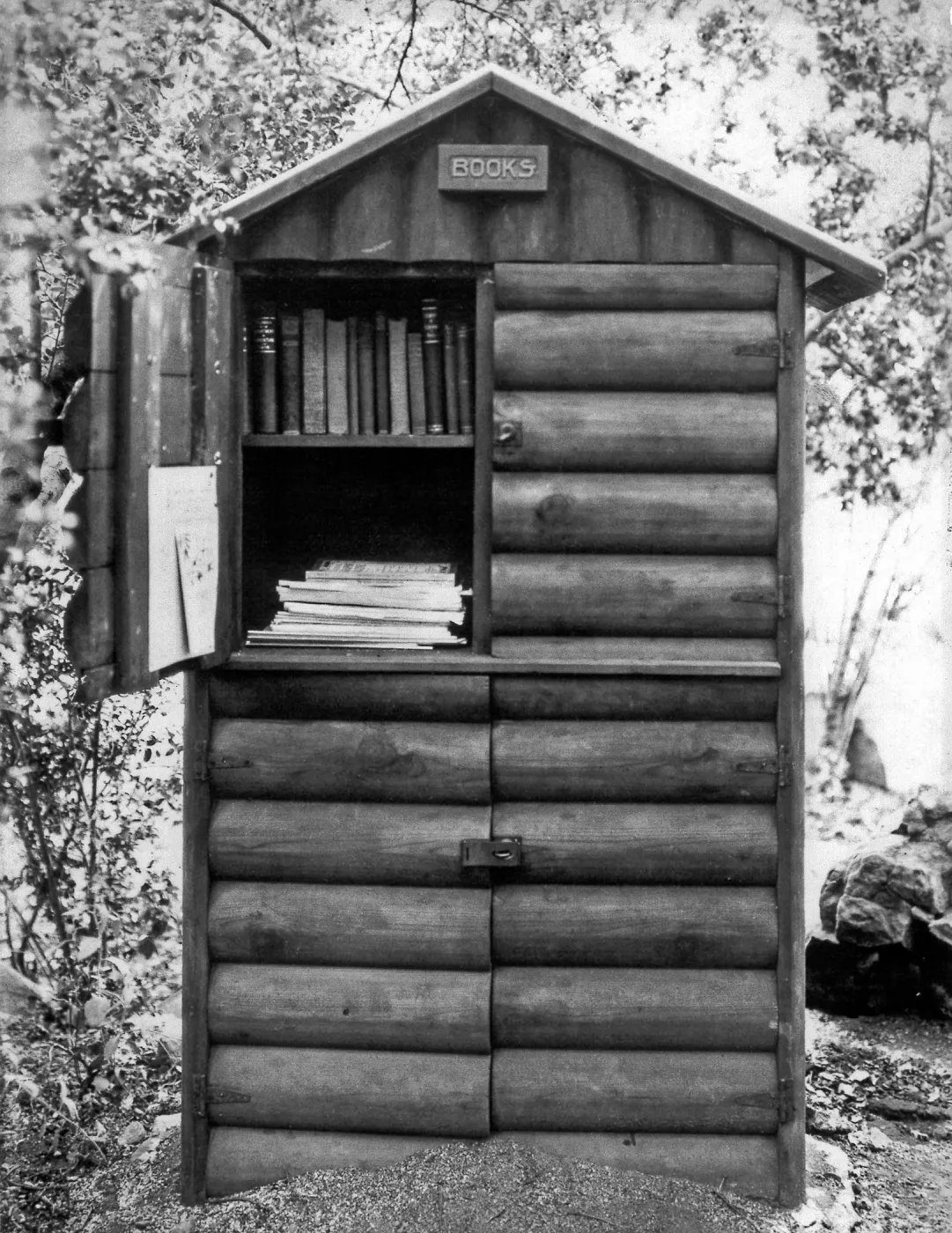 Black and white photograph of log cabinet labeled books with one door open and books inside in wooded area. 