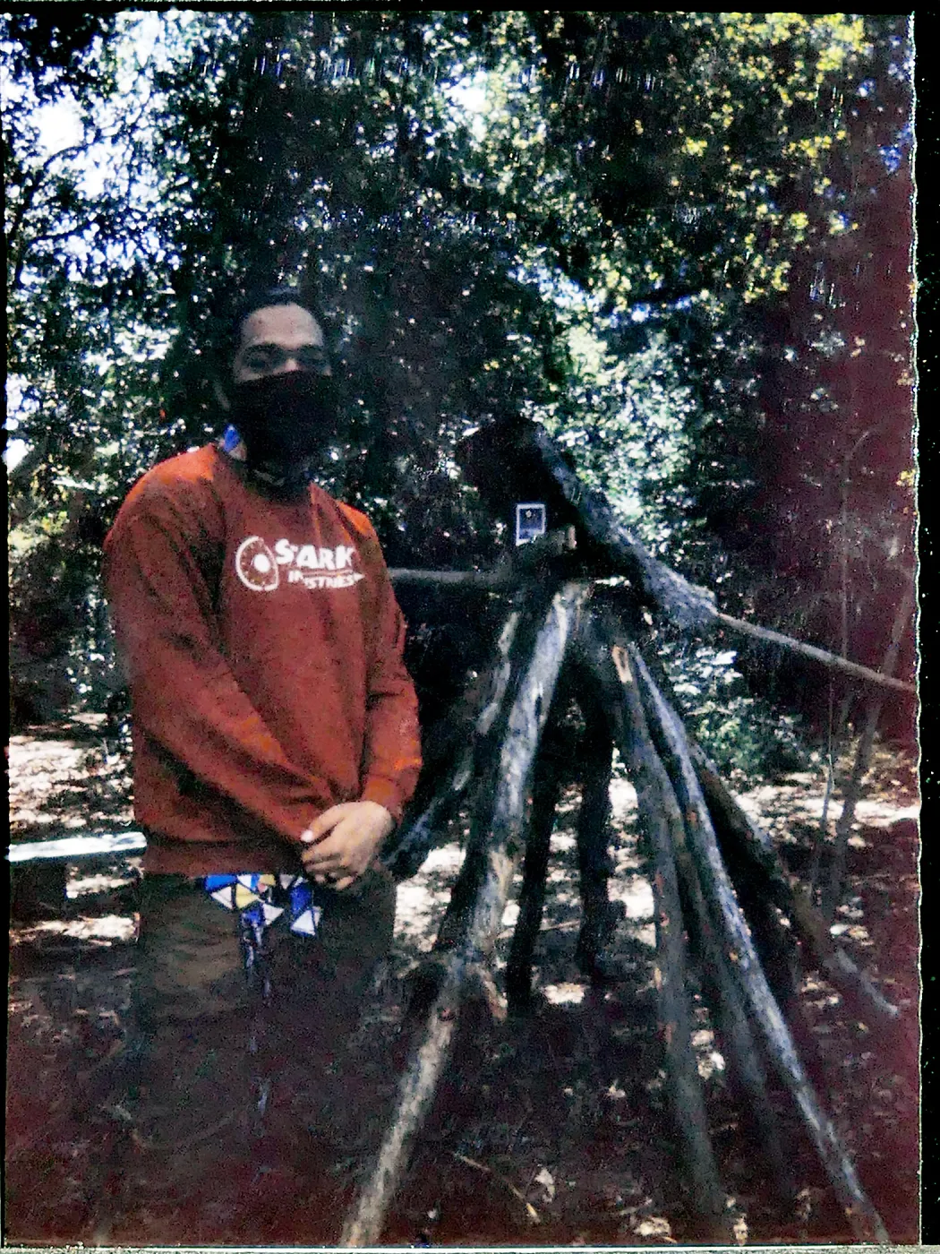Colored photo of a person wearing medical mask standing next to construction of large sticks in woods. 