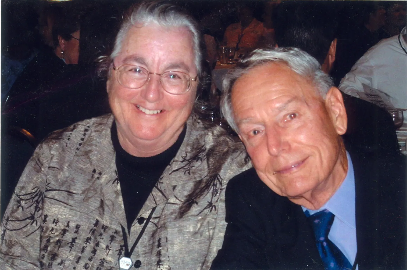 Flash photograph of female wearing black grey shirt with glasses and male wearing black suit blue tie both at event smiling. 