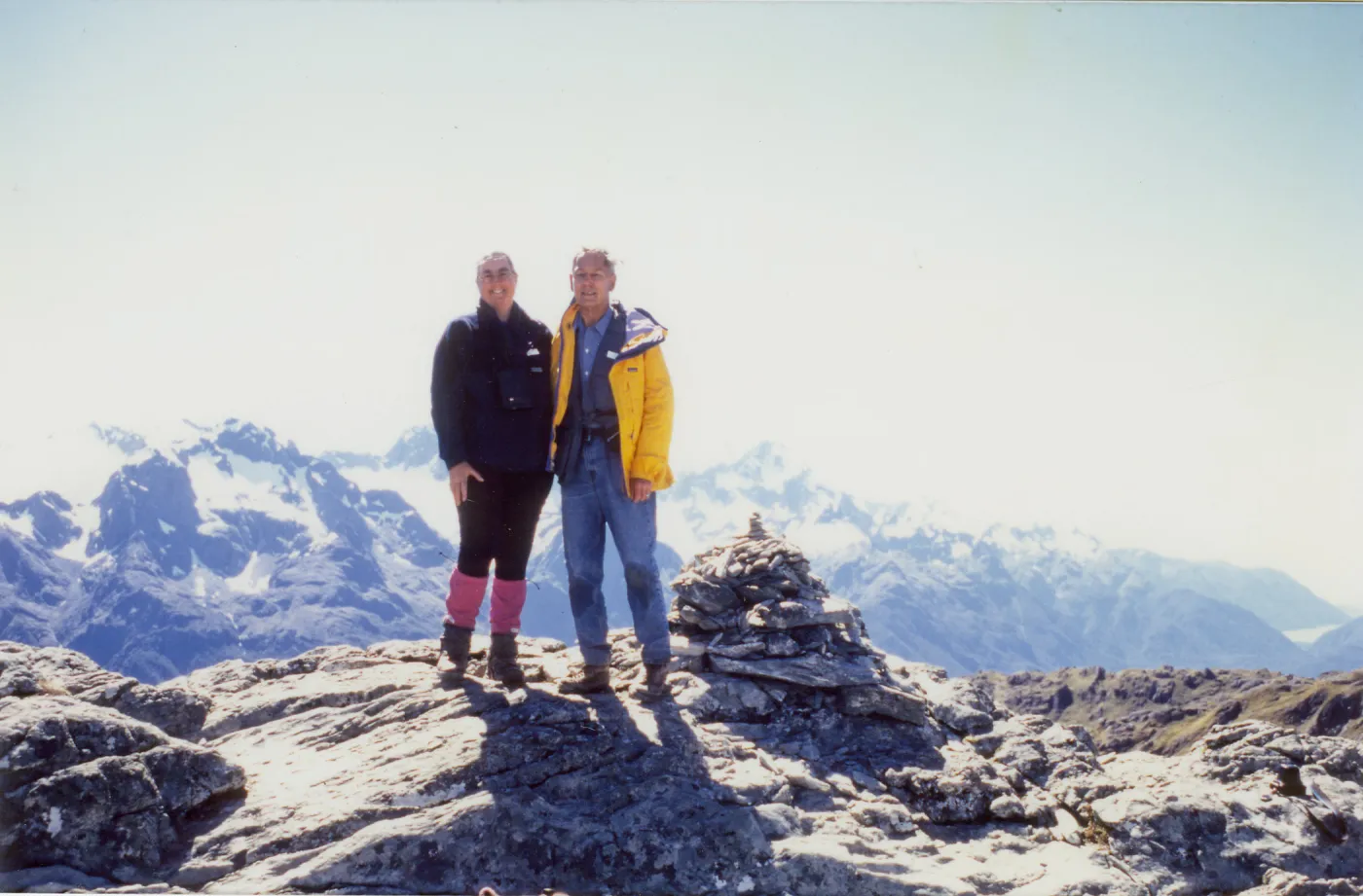 Photograph of male wearing yellow jacket and female wearing black and pink clothing standing on top of mountains with rocky and snowy terrain.