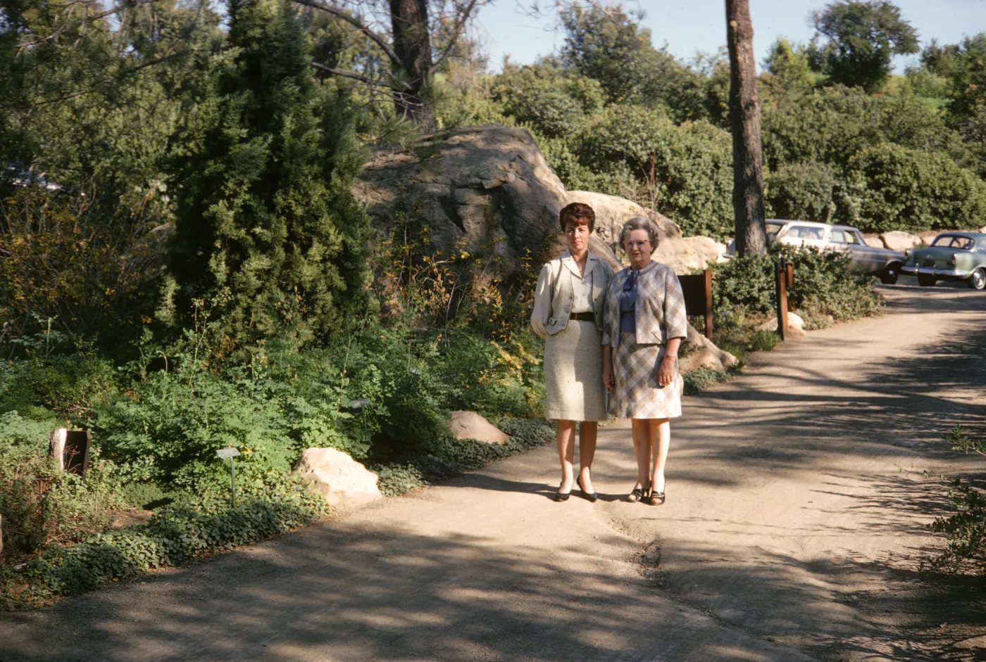 Two women with serious expressions wearing pale colored suits stand on an uneven paved road. Next to them at the edge of the road there is an evergreen tree among thick greenery, and behind them are a few large boulders and 2 pale colored, older model cars. 