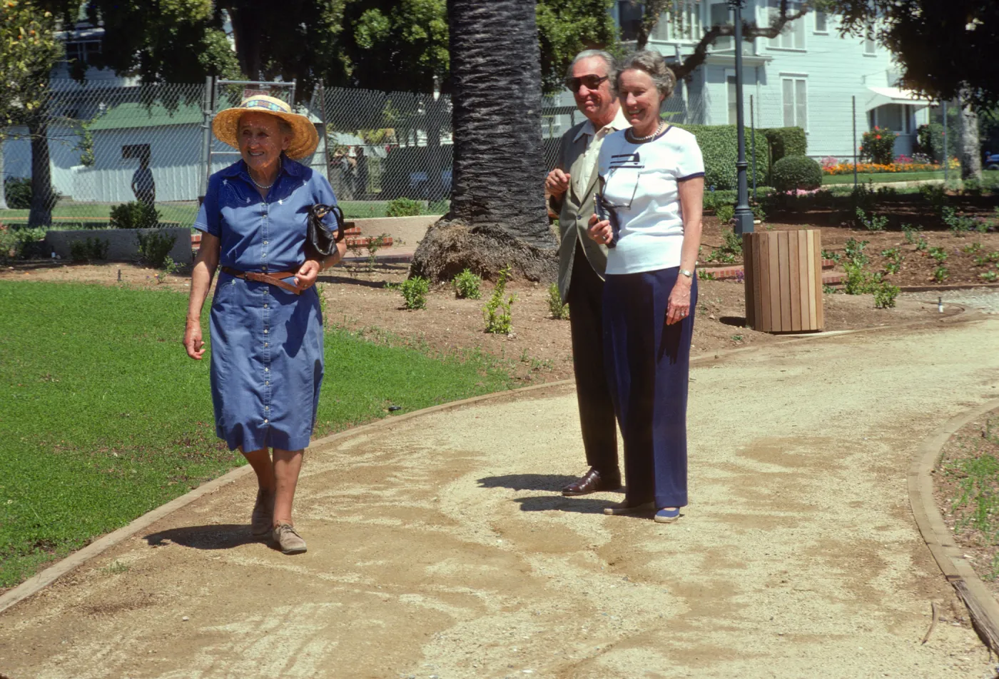 Three older people, a woman in a blue dress and straw hat, a woman in a white shirt and navy slacks, and a man in am olive suit and sunglasses, smile on a path between grass fields and large palm trees. 