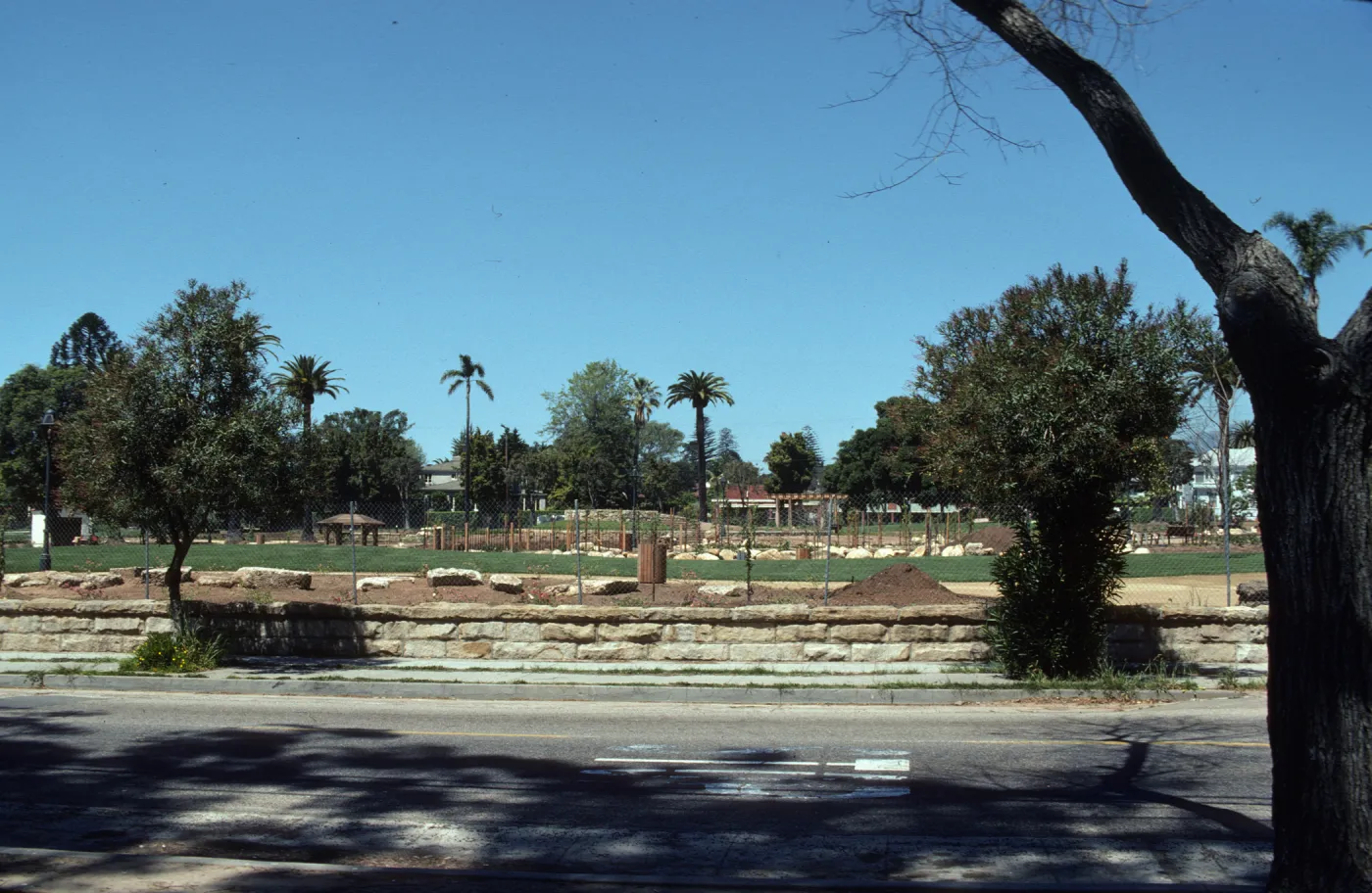 A park consisting of grassy areas and beds of soil dotted with large stones separated by paths viewed from across a paved street. 