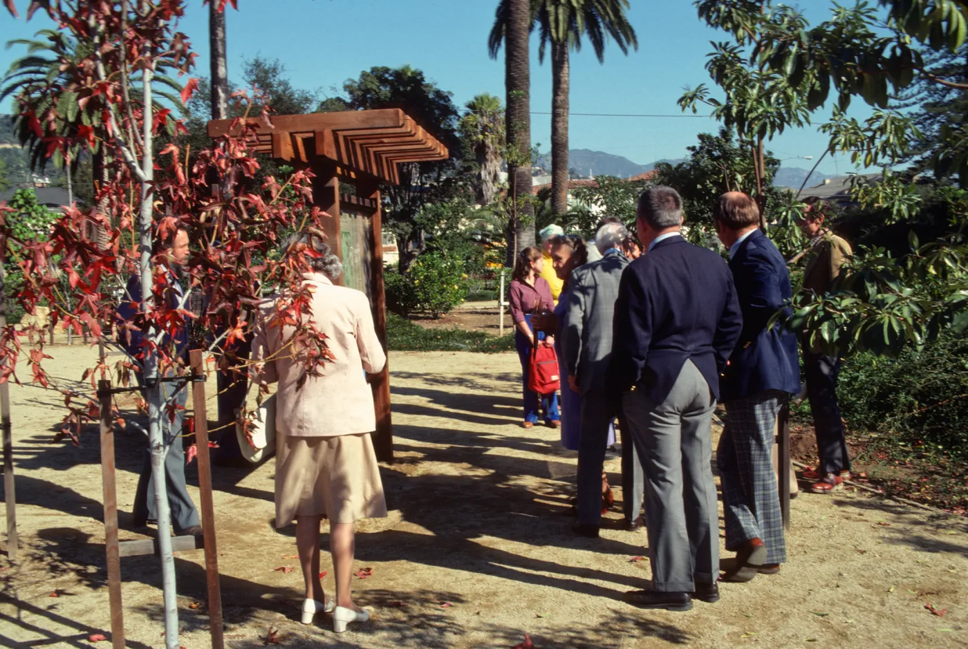 A group of well-dressed people gather in a garden around a large, wooden exhibit sign containing a map. 