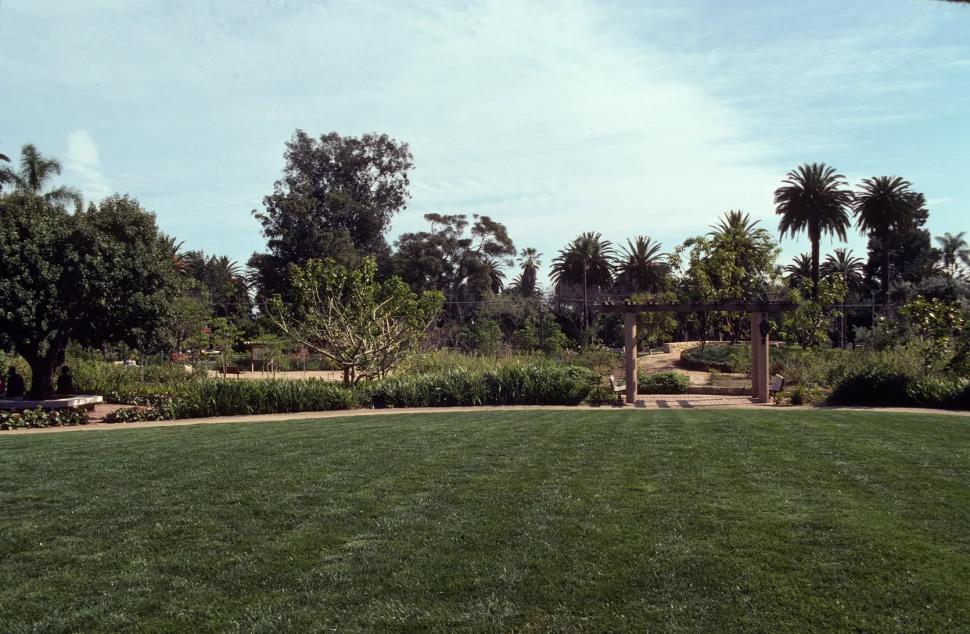 A uniform, grassy yard bordered by a walking path, several trees and bushes, and two benches beneath a pergola. 
