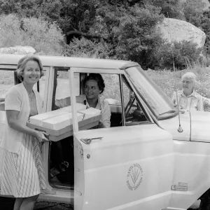 a black and white photo of three women by a parked car labeled one woman stands outside holding a cooler, another sits in the driver's seat, and an older woman stands behind the vehicle in a tree-lined area.