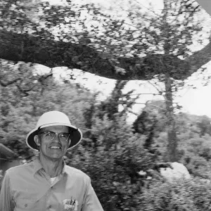Black-and-white photo of a smiling man in glasses and a brimmed hat, wearing a button-up shirt, standing outdoors with trees and shrubs behind him.