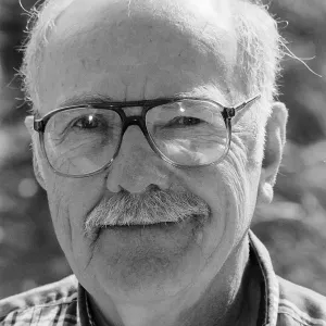 Black and white close-up of an older man wearing glasses and a plaid shirt, looking at the camera.