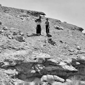 Black and white photograph of three people on a rocky hillside, two of which are underneath a black umbrella.