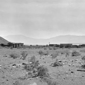 Black and white photograph of an abandoned town of wooden buildings with mountains in the background.