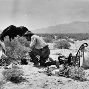 Black and white photograph of a man in a hat kneeling in a desert landscape next to a pile of equipment with a black horse grazing behind him.